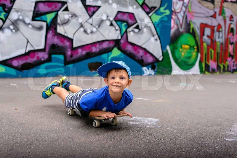 Small boy lying on his skateboard using his hands to propel himself along in front of a colorful painted graffiti wall, close up with him facing the camera, stock photo