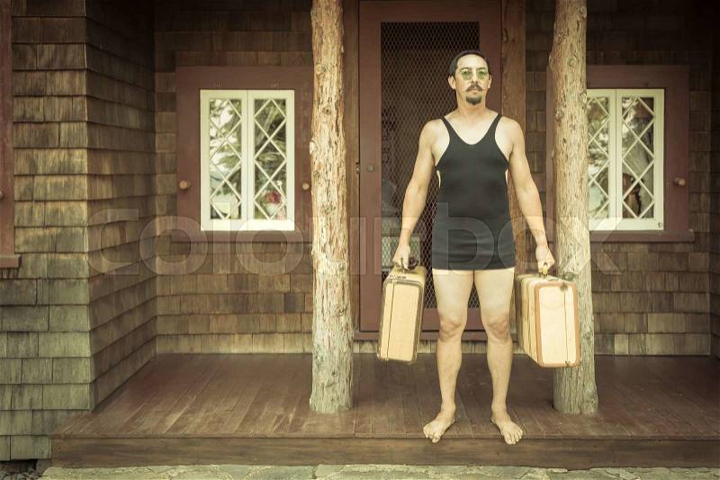 Goofy Gentleman Dressed in 1920’s Era Swimsuit Holding Suitcases on Porch of Cabin, stock photo
