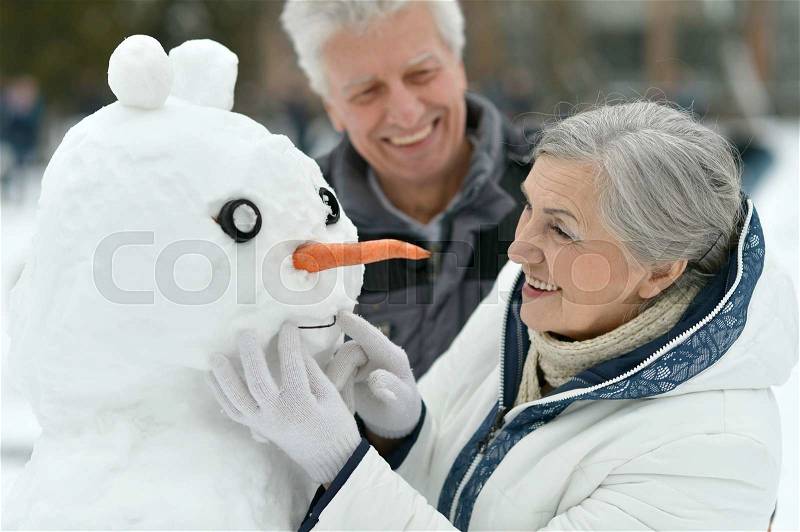 Happy senior couple with snowman in winter outdoors, stock photo