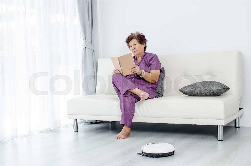 Senior woman reading a book while robot vacuum cleaning floor at home. Modern lifestyle concept, stock photo