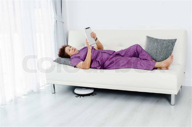 Senior woman unsing tablet while robot vacuum cleaning floor at home. Modern lifestyle concept, stock photo
