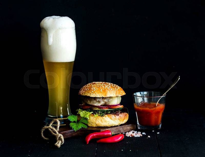 Fresh homemade burger on wooden serving board with spicy tomato sauce, sea salt, herbs and glass of light beer over black background, stock photo