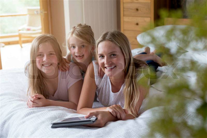 Sisters lying on a bed in their home with a digital tablet, smmiling for the camera, stock photo