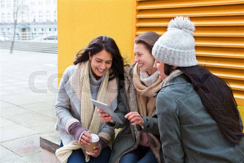 Three young women sitting in the city, looking at something on a digital tablet, stock photo