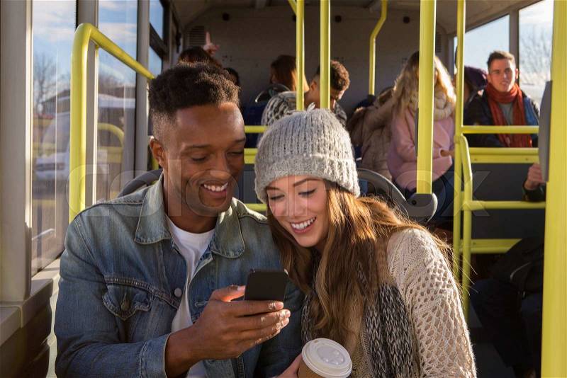 Young couple on a bus. They are both looking at something on a smartphone and smiling. There are people in the background who are also on the bus, stock photo