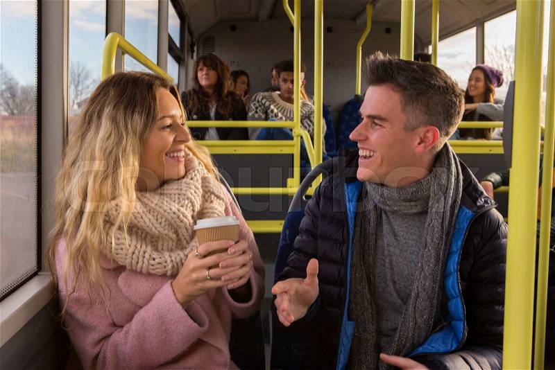 Young couple/friends chatting on a bus together. There are other people on the bus in the background, stock photo