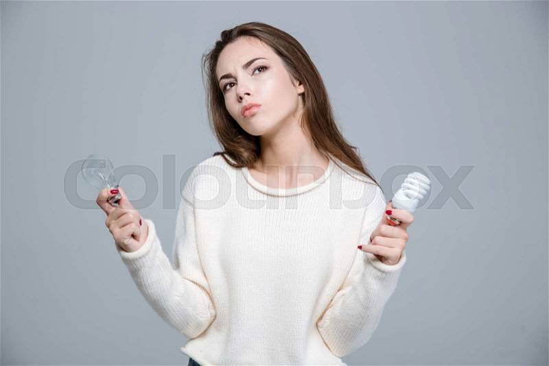 Portrait of a thoughtful woman holding saving light bulb and normal light bulb over gray background, stock photo