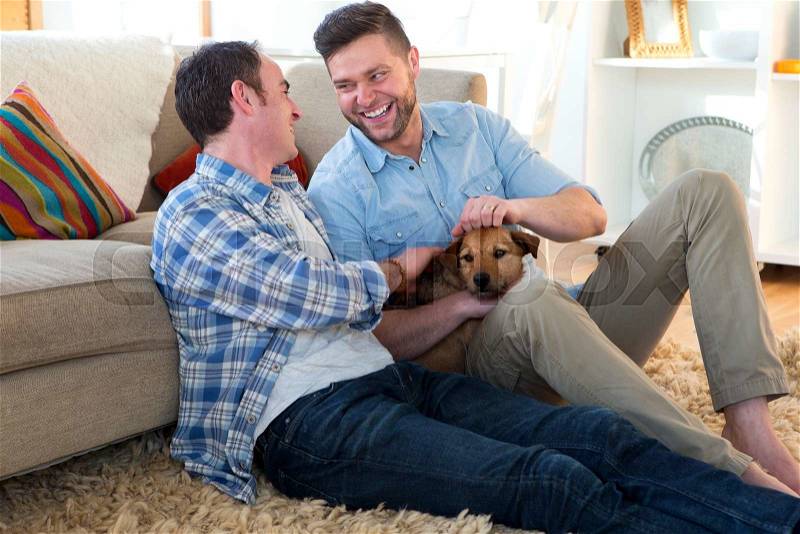 Same sex male couple sitting on the floor in their living room with their dog, stock photo