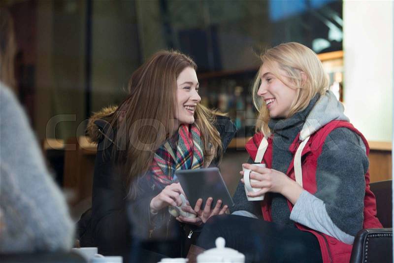 Two women are sitting in a cafe. They are talking and looking at a digital tablet with coffees in their hand, stock photo
