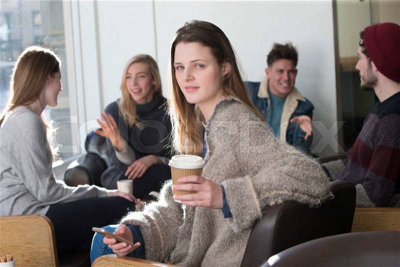 Young woman is smiling for the camera in a cafe with her friends in the background. She is holding a smartphone and a cup of coffee, stock photo