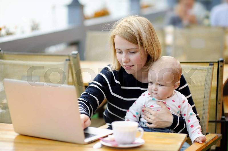 Young mother with her adorable baby girl working or studying on her laptop in outdoor cafe, stock photo