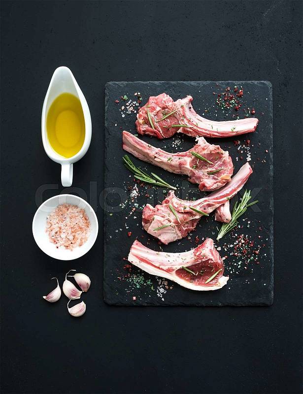Raw lamb chops. Rack of Lamb with garlic, rosemary and spices on black slate tray, oil in a saucer, salt, dinnerware over dark rustic wood background, stock photo