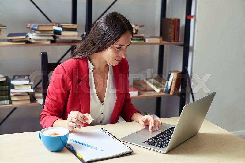 Young woman working with laptop and eating cookies in office, stock photo