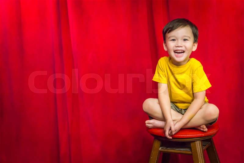 Laughing Mixed Race Boy Sitting on Stool in Front of Red Curtain, stock photo