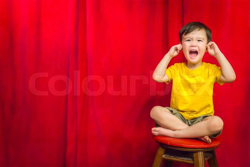 Mixed Race Boy With His Fingers In His Ears Sitting on Stool in Front of Red Curtain, stock photo