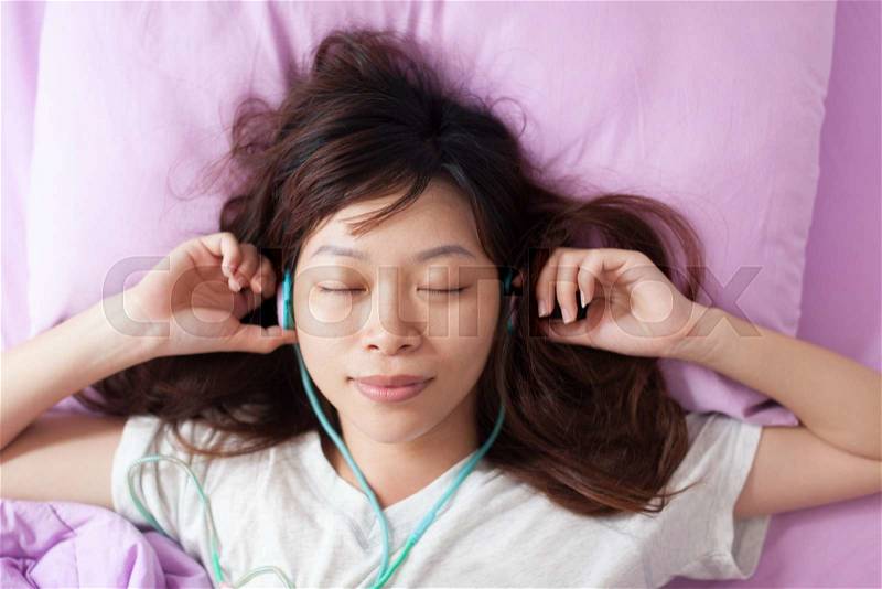 Asian young girl listen to music closed eyes wear headphones lying on bed top angle view, stock photo