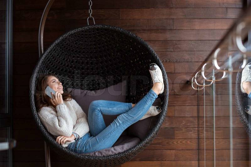 Beautiful happy girl weared in white sweater and blue jeans lying in a bubble chair and talking on the cellphone, stock photo