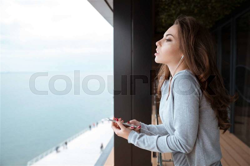 Attractive young woman with beautiful long hair standing on the balcony and listening to music, stock photo