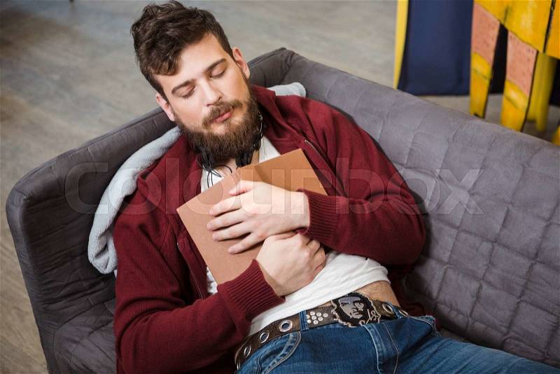 Tired handsome young guy with beard sleeping hugging book on grey sofa, stock photo