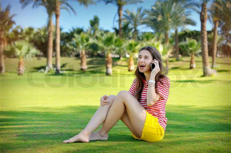 Beautiful young woman sitting on a green lawn and talking on a cell phone, stock photo