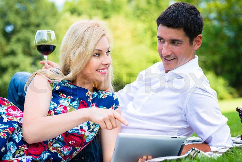 Couple at picnic in park with red wine and tablet computer, stock photo