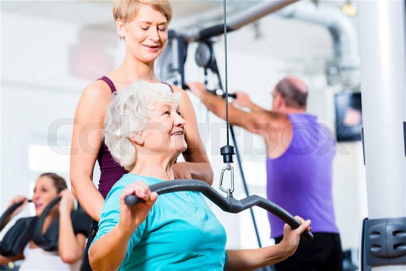 Senior woman doing back training with trainer in gym at machine, stock photo