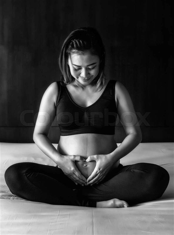 Pregnant woman holding her hands on her swollen belly shaping a heart, toned black and white, stock photo