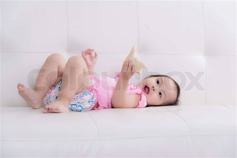 Cute Asian baby lying on sofa and eating bread at home, stock photo