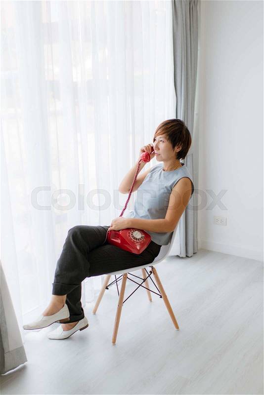 Smart Asian woman sitting and holding red vintage phone near window at home, vintage toning, stock photo
