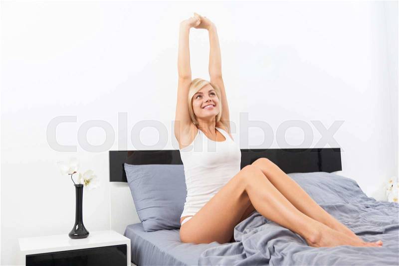 Woman wake stretching raised arms hands up, young girl sitting on her bright bed at home, stock photo