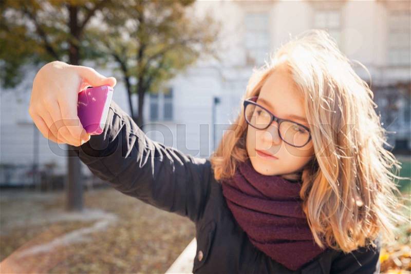 Beautiful Caucasian blond teenage girl taking photo on her smartphone in autumnal park, selective focus on hand with phone, shallow DOF, stock photo
