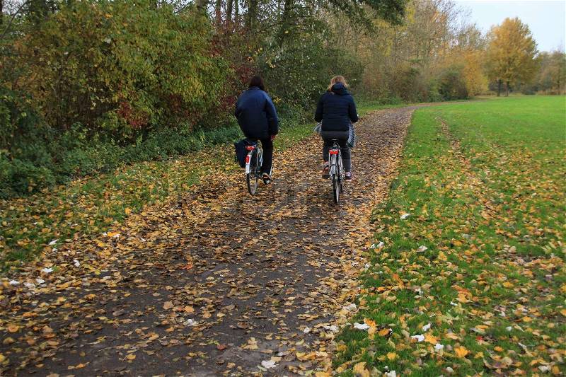 Two ladies are cycling through the fallen leaves on the path in the park at the country side in autumn, stock photo