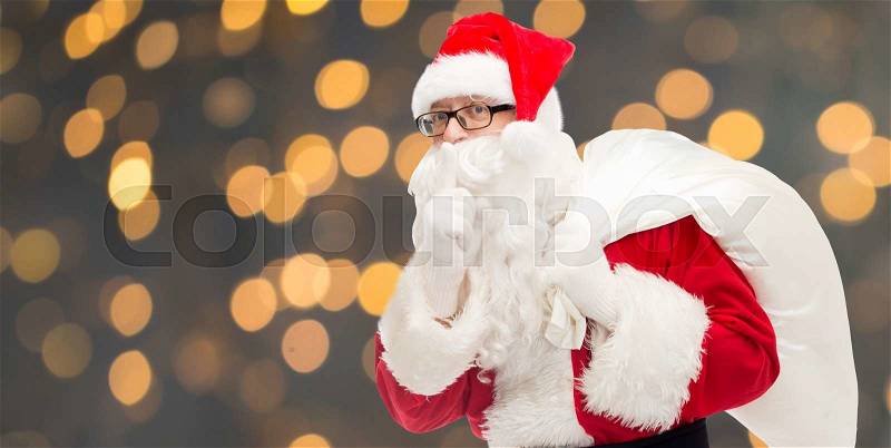 Christmas, holidays and people concept - man in costume of santa claus with bag making hush gesture over golden lights background, stock photo
