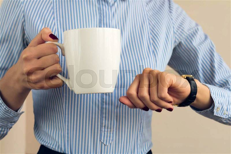 Woman with a burgundy manicure in a blue striped shirt holds a white cup and looking at her watch, stock photo