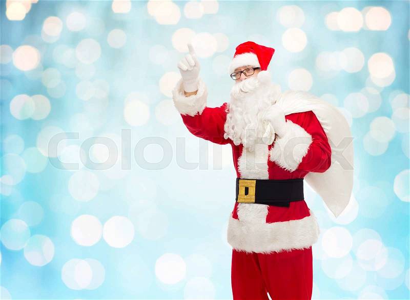 Christmas, holidays, gesture and people concept - man in costume of santa claus with bag pointing finger up over blue holidays lights background, stock photo