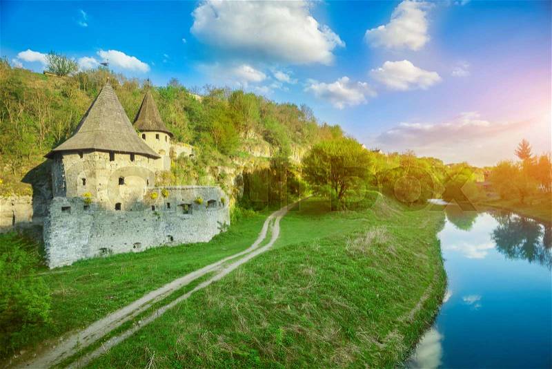 Ancient stone castle in Kamianets Podilskyi standing on hill with green trees and bushes with rural road and river, stock photo