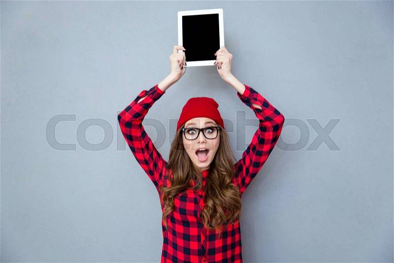 Portrait of a cheerful hipster woman showing blank tablet computer screen over head on gray background, stock photo