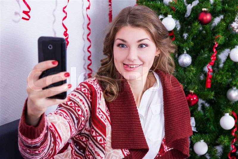 Portrait of funny young woman taking selfie photo with mobile phone near christmas tree, stock photo