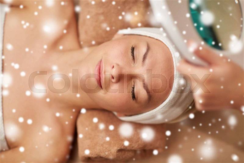People, beauty, skin care, winter and relaxation concept - close up of beautiful young woman lying with closed eyes and beautician looking through magnifying lamp in spa salon with snow effect, stock photo