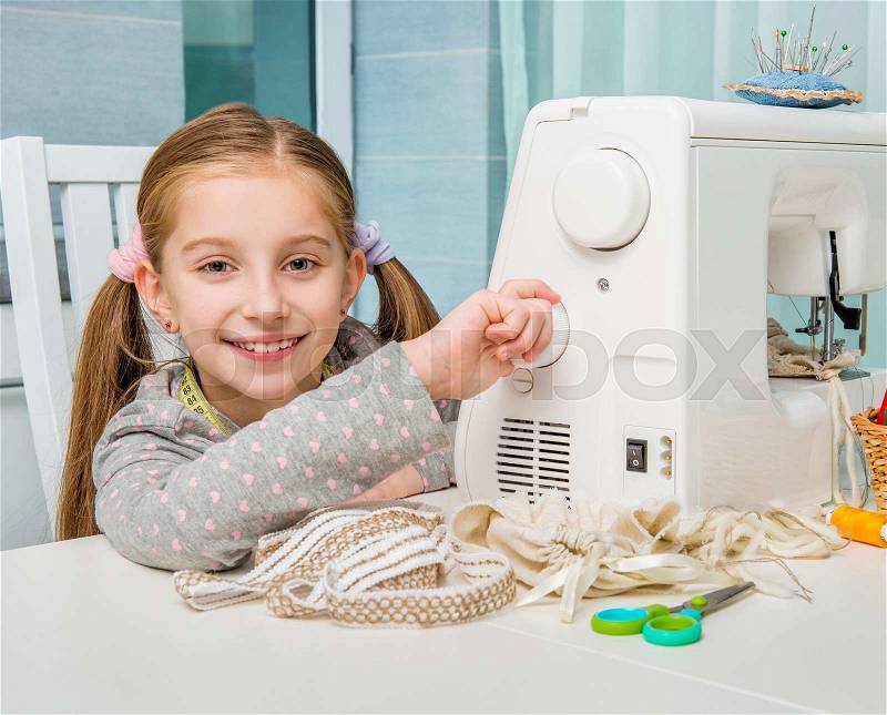 Smiling little girl at the table with sewing machine looking at camera, stock photo