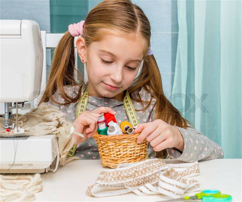 Little seamstress sitting at the table with threads in basket and sewing machine, stock photo