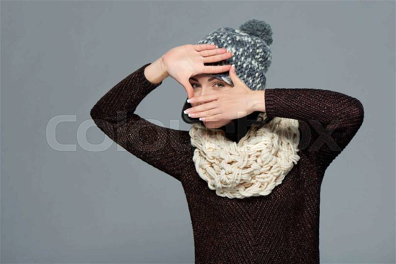 Pretty young woman in winter clothing making a frame with her hands, over grey background, stock photo