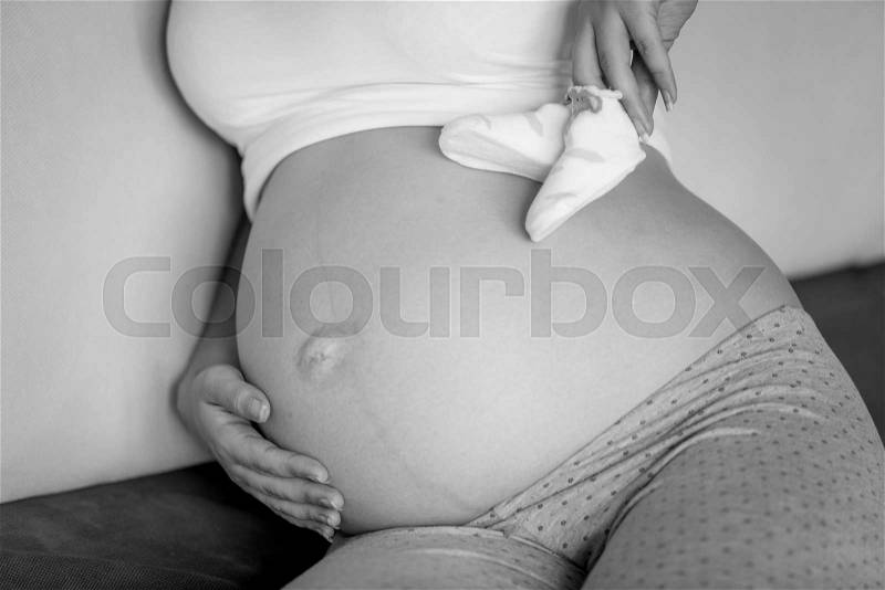 Closeup black and white photo of pregnant woman posing with baby socks on belly, stock photo