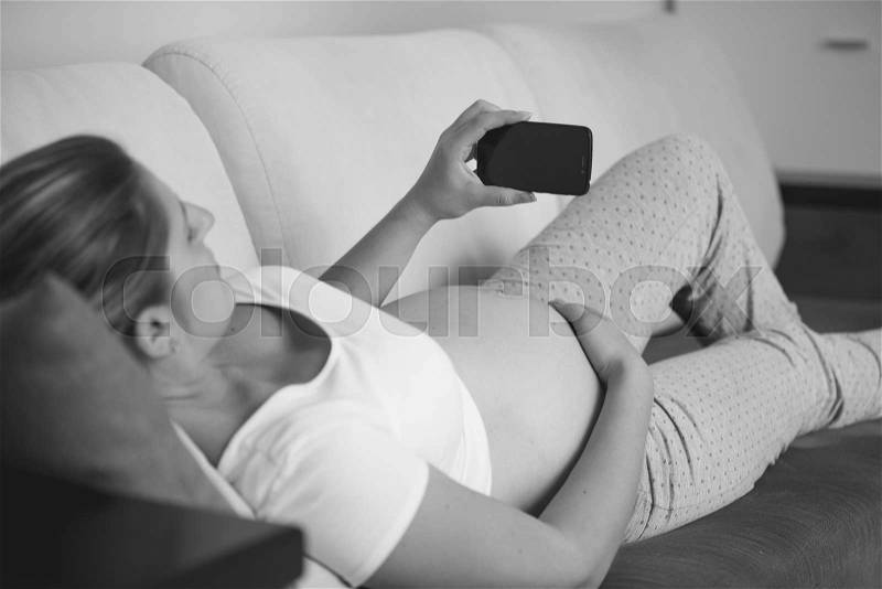 Closeup black and white shot of pregnant woman relaxing on sofa and using mobile phone, stock photo