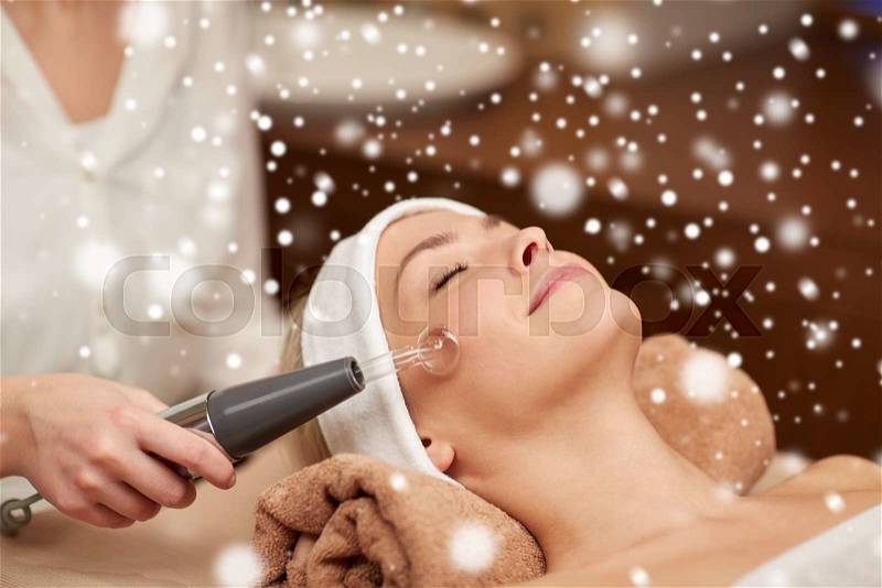 People, beauty, spa, cosmetology and technology concept - close up of beautiful young woman lying with closed eyes having face massage by massager and beautician in spa with snow effect, stock photo