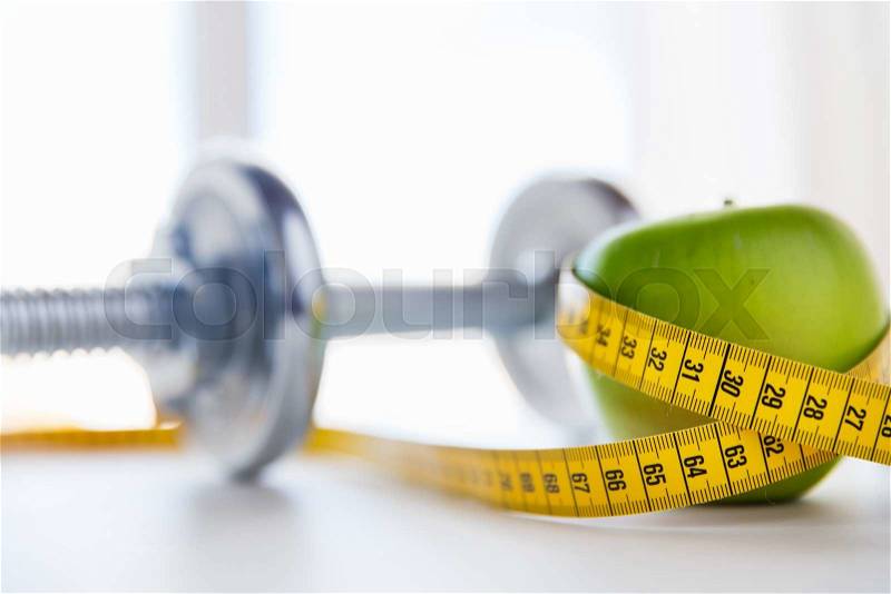 Sport, fitness, diet and objects concept - close up of dumbbell and green apple with measuring tape, stock photo