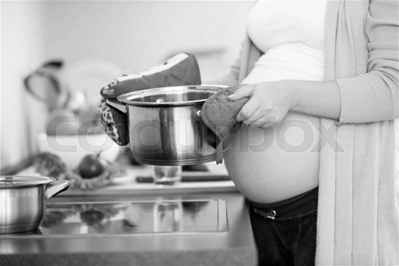 Black and white closeup photo of pregnant woman holding metal pan on kitchen, stock photo