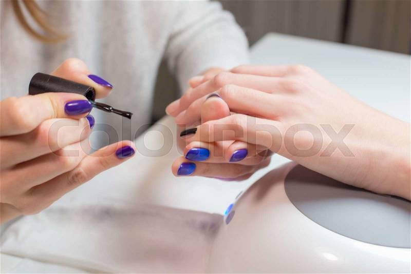 Beautician paints nails woman client in the spa salon. The process takes place at white table near professional tools and Uv lamp, stock photo