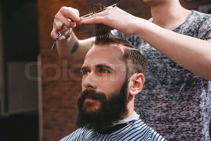 Portrait of young handsome man with beard having a haircut with comb and scissors in hair salon, stock photo