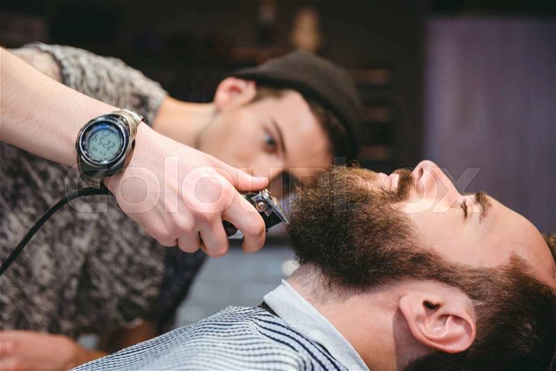 Attractive bearded man getting his beard shaved by skillful barber in barber shop, stock photo